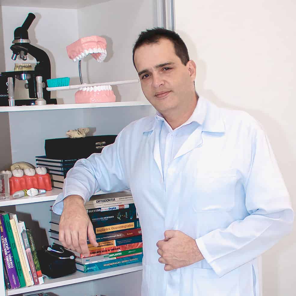 Doutor Rafael Muglia, Dentista especialista em Implantodontia com avental branco e discreto sorriso em frente a uma estante com livros, microscópio, modelos didáticos. É um homem de 45 anos de descendência italiana, já um pouco calvo e não muito magro.